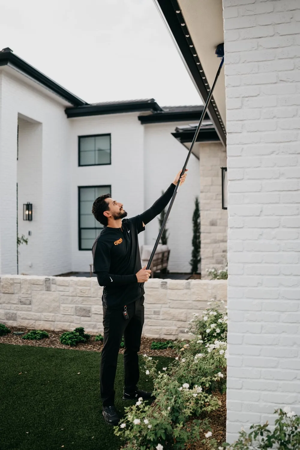 Citrus Pest Co technician reaching up with an extension duster to clear webs from the eaves of a white brick East Valley home
